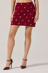 A woman is wearing a high-waisted, burgundy mini skirt decorated with small, white flower patterns scattered throughout. She is also wearing burgundy high-heeled shoes with ankle straps.